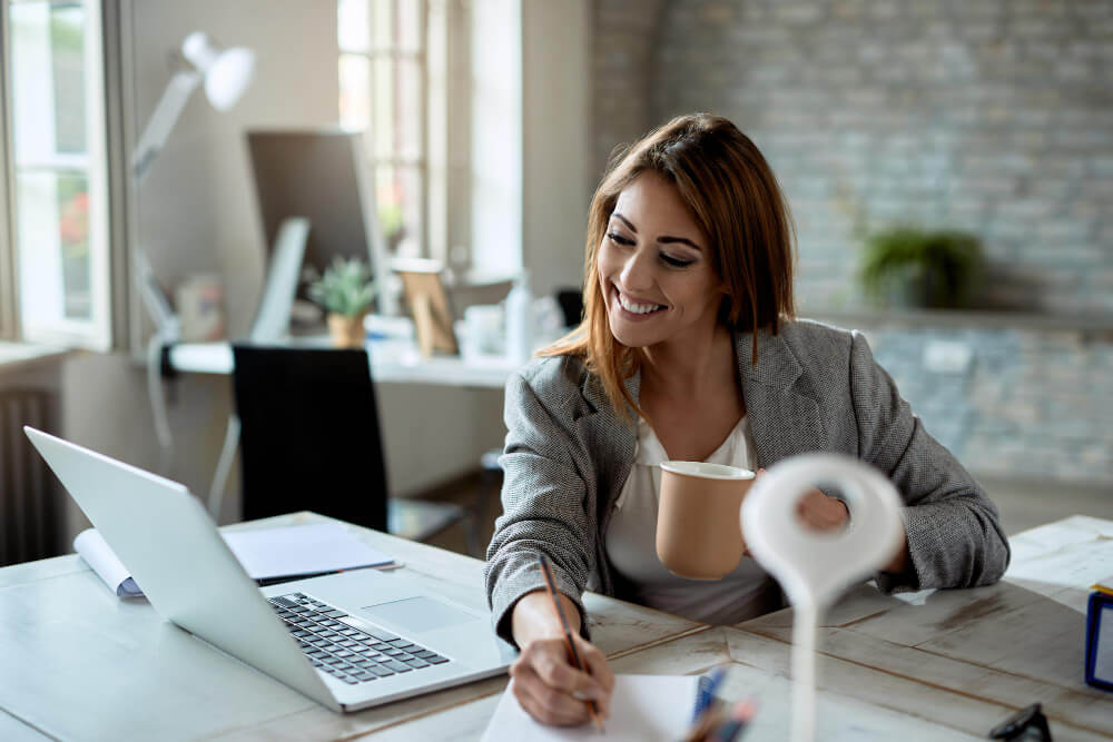 young-happy-female-entrepreneur-using-computer-taking-notes-while-drinking-coffee-office (1) middle bed