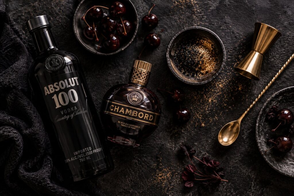 Black magic martini ingredients laid out on dark stone: black vodka, blackberry liqueur, edible black glitter, and dark cherries with a jigger and bar spoon